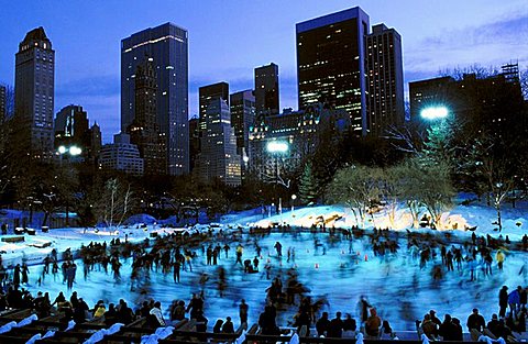 Twilight view of the Wollman ice skating rink in Central Park, New York City with buildings in background and skaters in blurred motion on the ice, January
